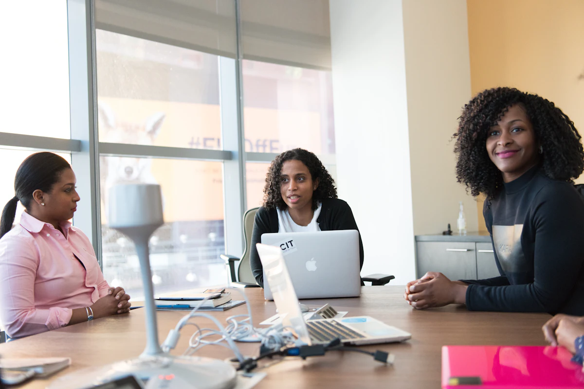 Three Black women in conversation around a table with a laptop in a bright office